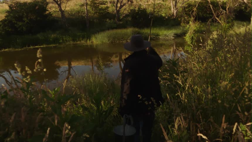 An elderly man with a fishing pole hikes down through tall grass to the water