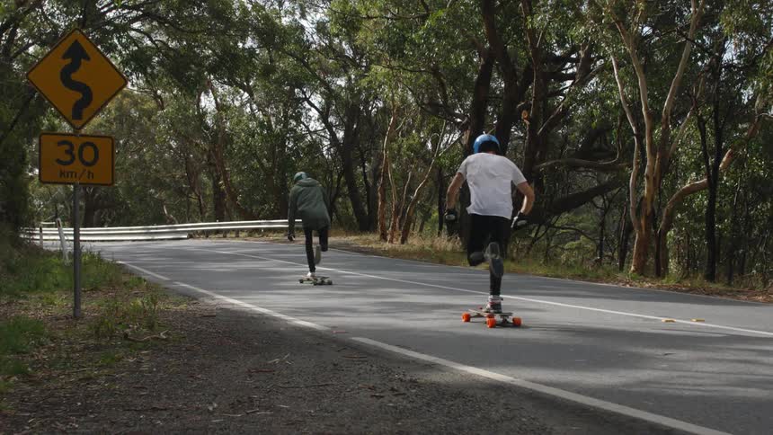 Two friends ride skateboards down a bend in the road