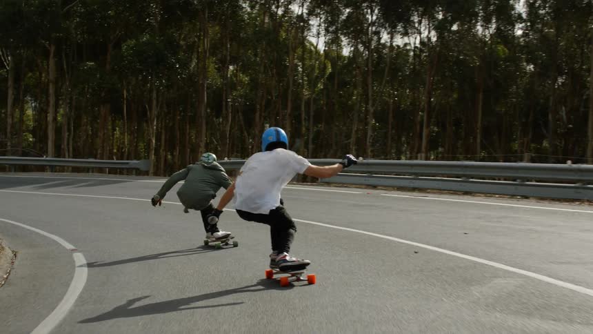 Two young people riding skateboards on a road downhill close to a forest