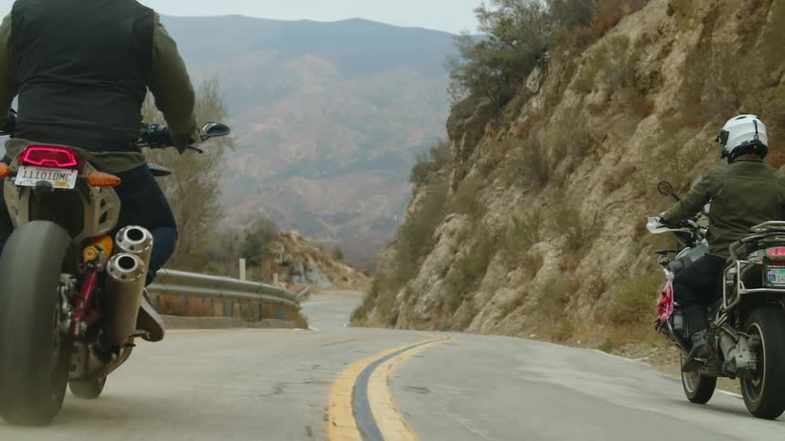 Two men drive motorcycles past hills