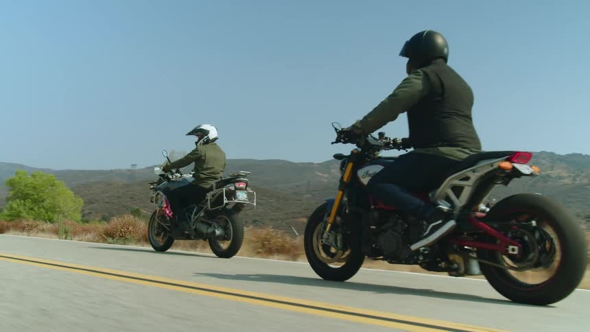 Two people are riding motorcycles on a road overlooking mountains under a clear sky
