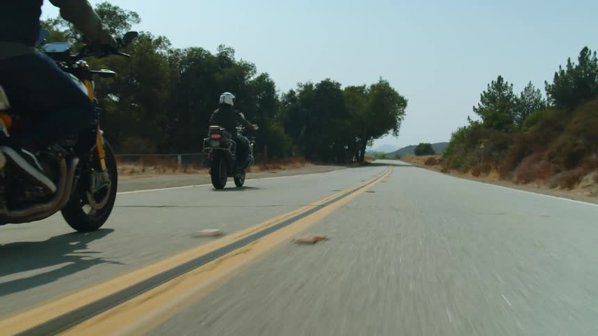 Two men drive motorcycles down a street next to a grassland