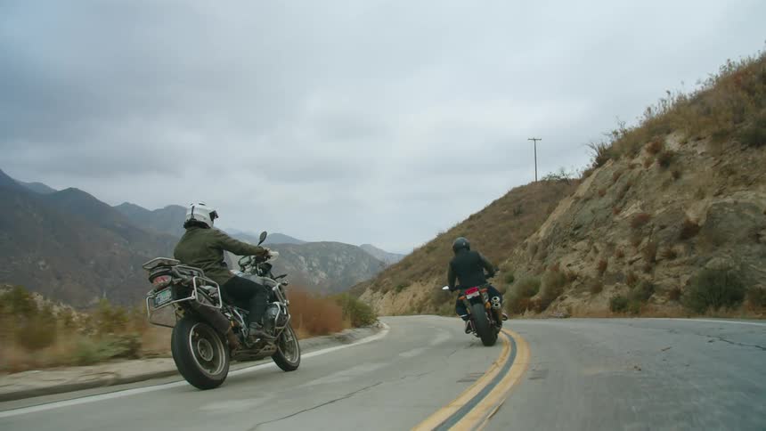 Motorcyclists ride motorcycles along highway between hills and drop into a valley on a foggy day