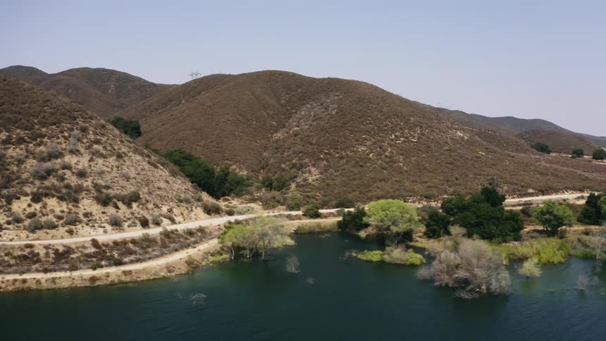 Two motorcycles traveling on a curving road at the base of hills next to a blue lake