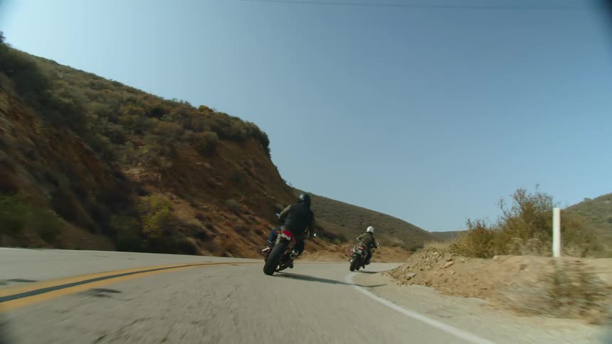 Two people drive motorcycles on a road next to hills