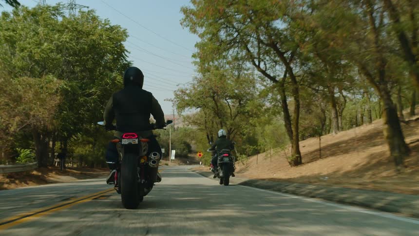 Two people are riding motorcycles on a road lined with trees and mountains in the distance