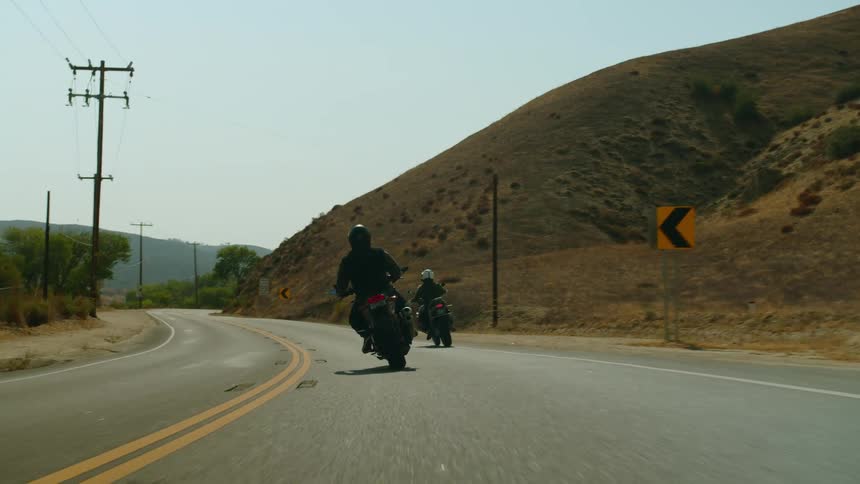Two people are riding motorcycles on a mountain road with mountains in the distance