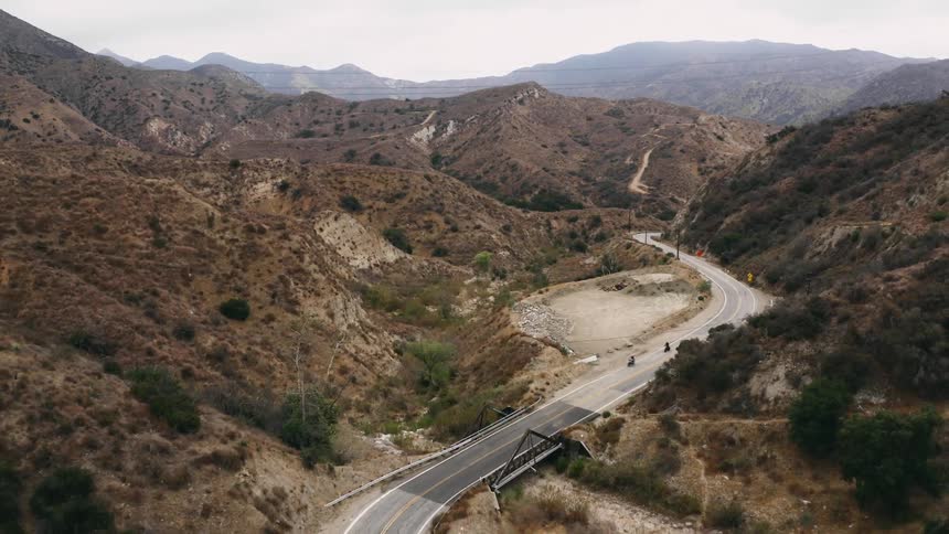 Two people drive motorcycles on a road through the hills