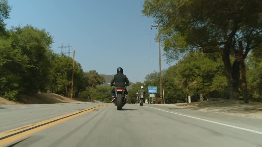 Two people are riding motorcycles on a road lined with trees and mountains in the distance