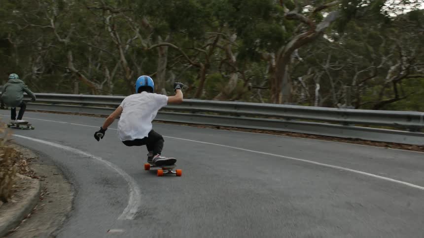 Two skateboarders ride skateboards around curve of road past woods