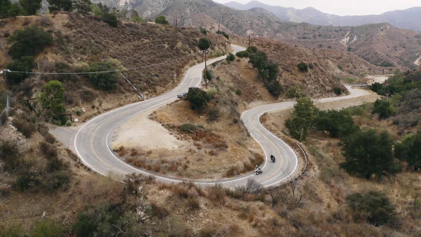 Two motorcyclists ride around curve on winding road between hills with foggy mountains in the background