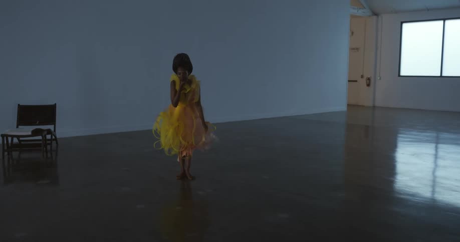 A woman wearing a yellow dress is bending down and she stands straight up and steps forward with bare feet
