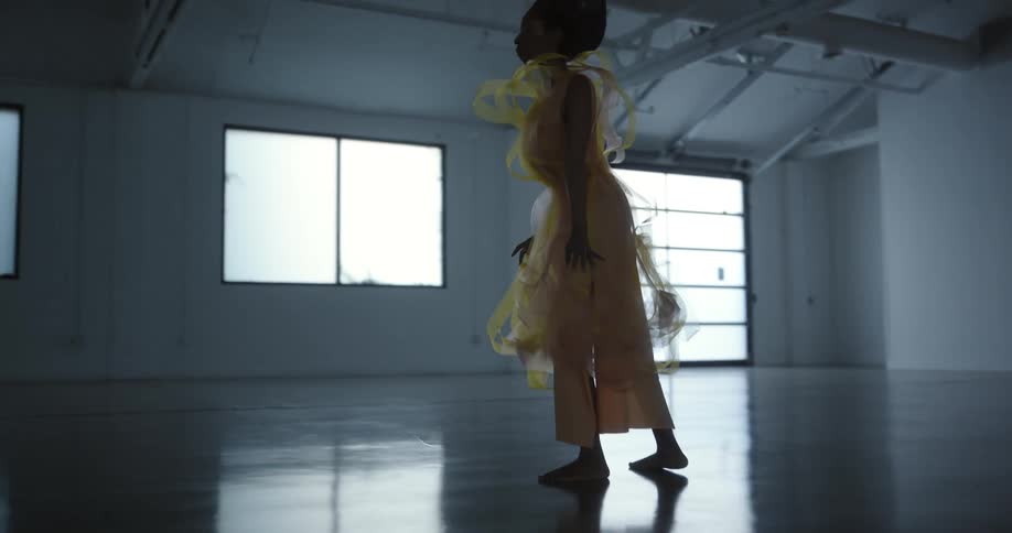 A woman wearing a yellow dress is standing barefoot on a shiny floor moving her arms in the air in a dance pose as light shines in from the windows