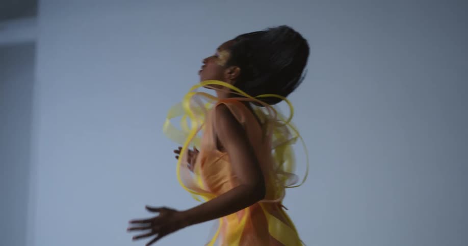 A woman in elaborate costume and hair dancing and reaching as she performs in a studio