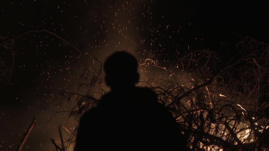 Man sits beside large bonfire as embers and smoke rise