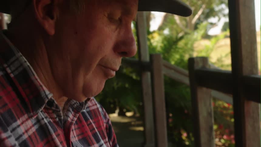 A man wearing a hat is standing on his porch looking down as the sun shines brightly on the trees