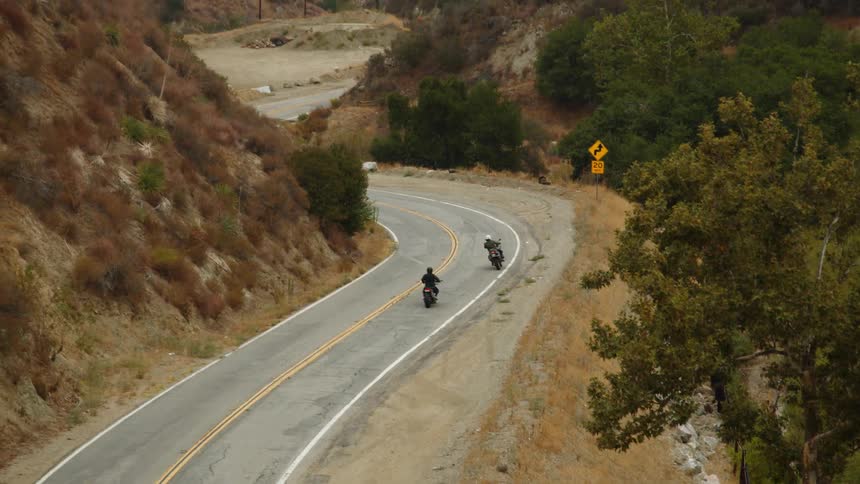 Two motorcyclists ride along highway between steep hill slope and drop off into forest