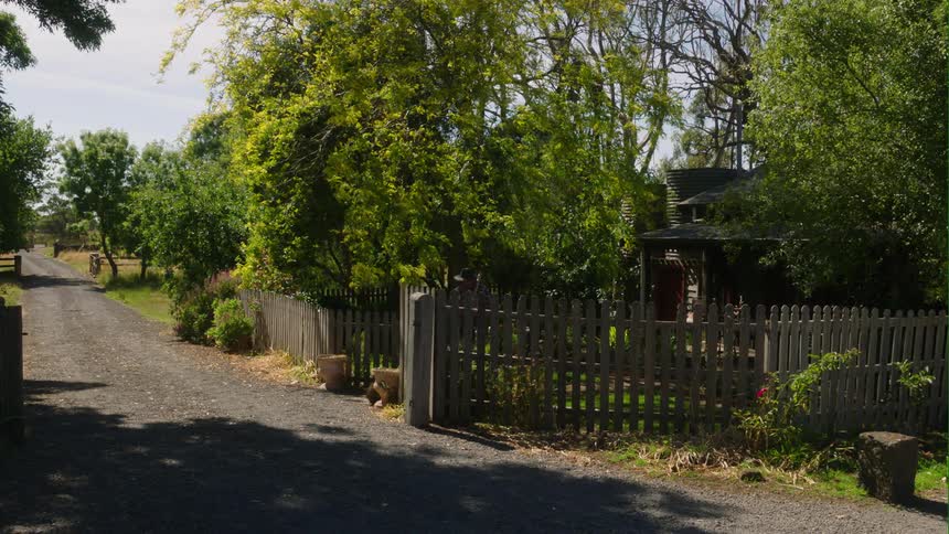 A man leaves his farm house and walks up the gravel road carrying a pickaxe on his shoulder