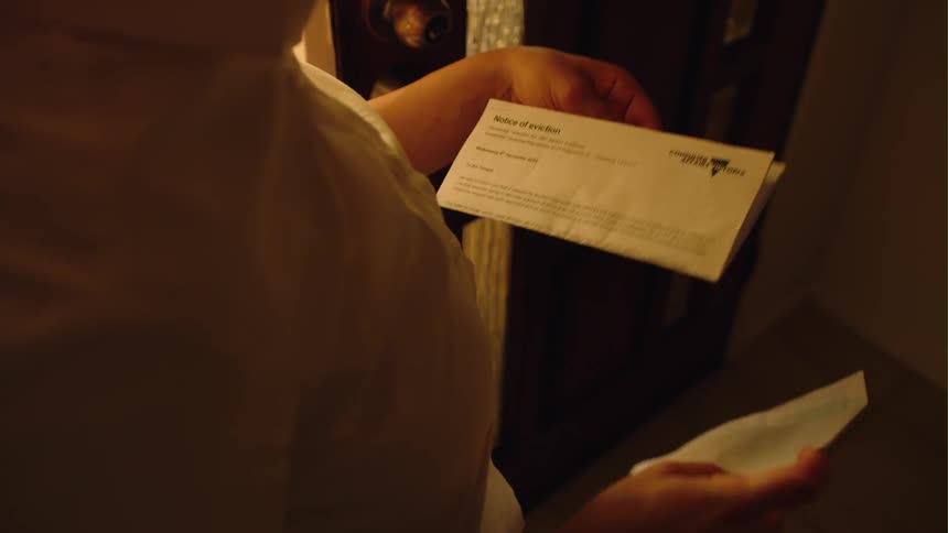 Mother stands near door as child plays on floor nearby and opens eviction letter