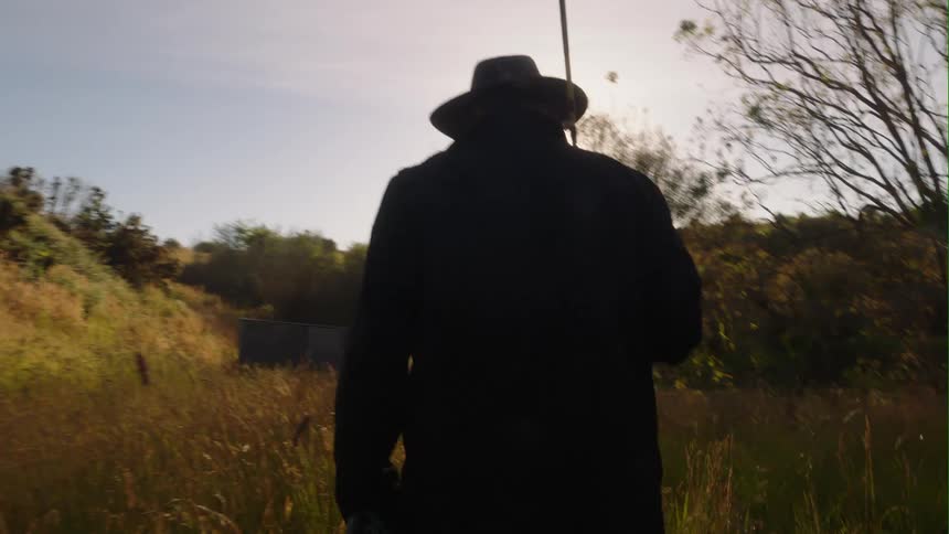 A man wearing a hat and a coat is walking through a field of tall grass and weeds near the woods carrying a fishing pole as the sun shines brightly upon him