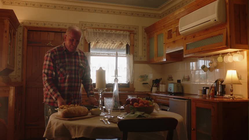 Older man places food in serving platters and bowls onto dining table set in kitchen