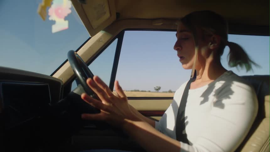A woman driving a vehicle wipes tears from her face and leans forwards to look at the dash