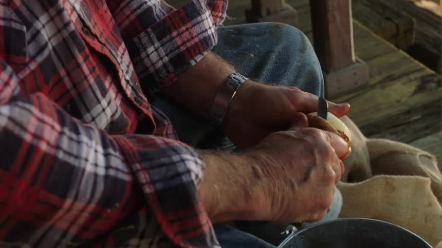 A man sitting on his porch peeling a potato using a pocket knife