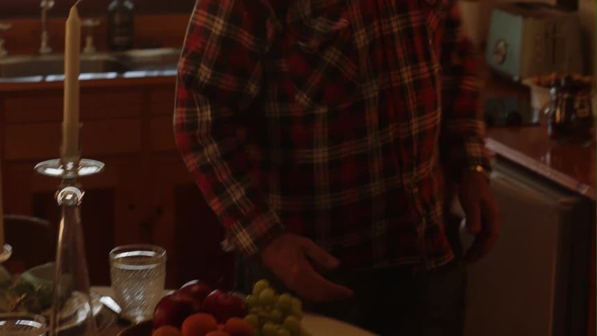 A man standing at the kitchen counter picks up a bowl of fresh fruit and places on the table