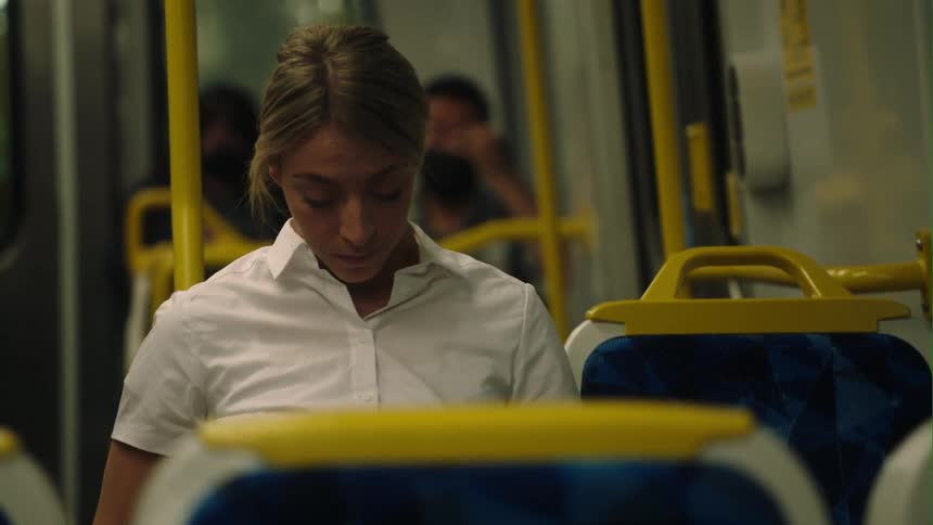 Blonde woman with ponytail and white shirt sits on tram looking down at smartphone