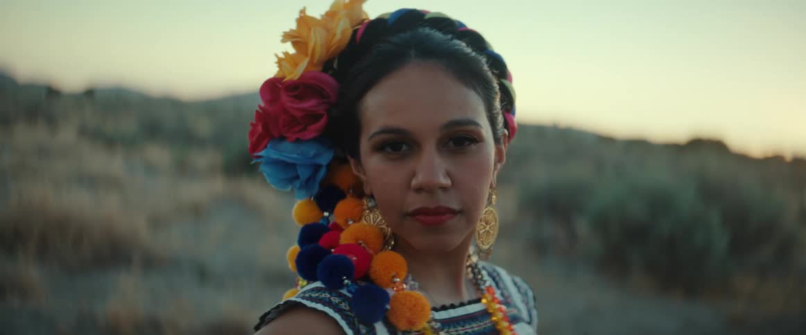 A woman with flowers in her hair stands in front of a hill