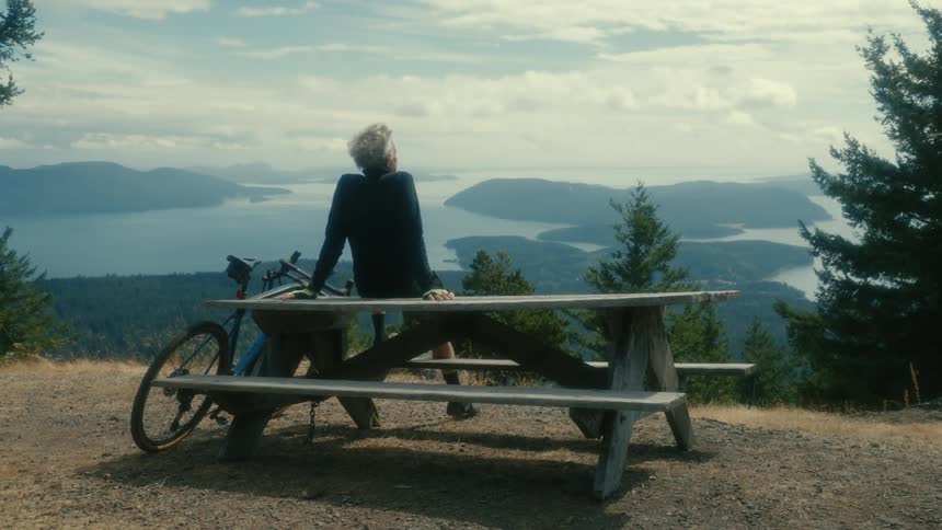 An elderly cyclist sits on a picnic bench next to his bike and takes in the great outdoors