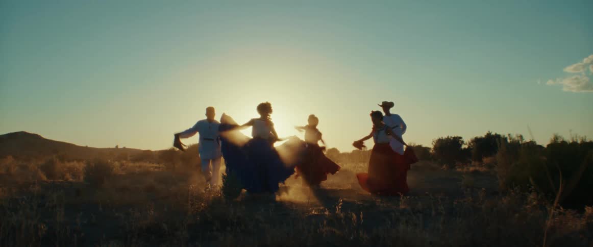 A group of people are outside dancing and the men are wearing cowboy hats and the women are wearing long skirts with flowers in their hair as the sun shines brightly upon them