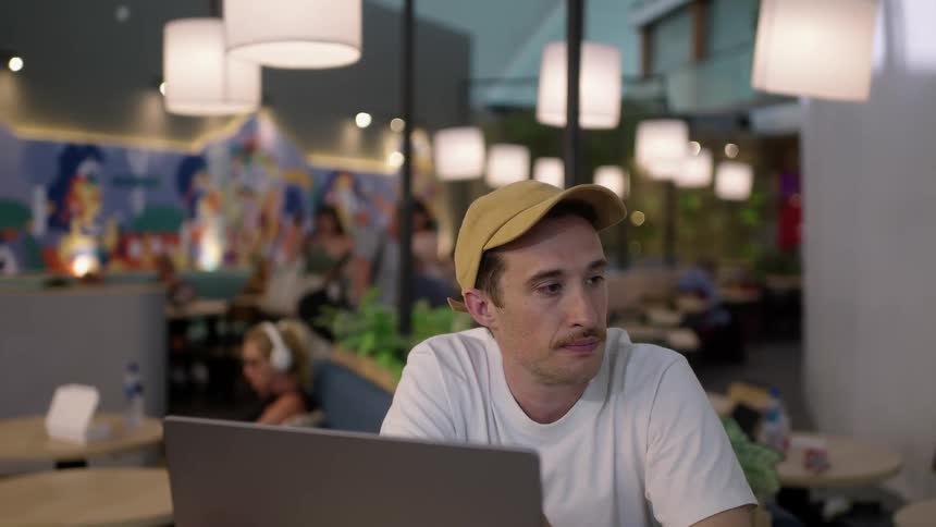A man is sitting at a table inside a cafe looking at his laptop computer