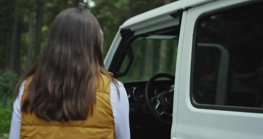 A woman walks to a white truck parked on a forest road and opens the door to get inside and holds the steering wheel as she shuts the door