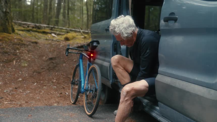 Older cyclist sits on floor of van parked near woods putting on shoes near bicycle