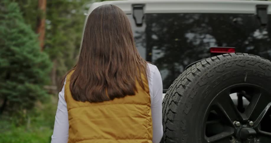 A woman parked in a forest gets out of her vehicle and goes to the back and gets a backpack out