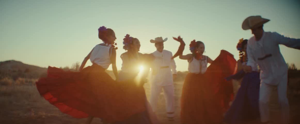 A group of people are outside dancing and the men are wearing cowboy hats and the women are wearing long skirts with flowers in their hair