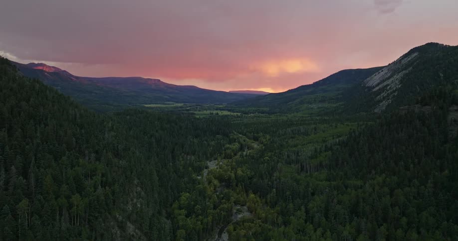 A narrow river winds through a forested mountain valley in the evening
