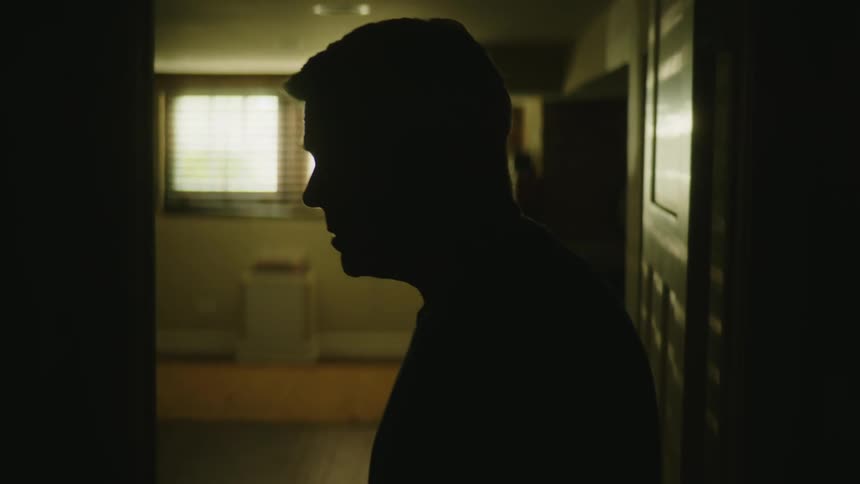 Man standing in doorway silhouetted against window light raises his head from looking down