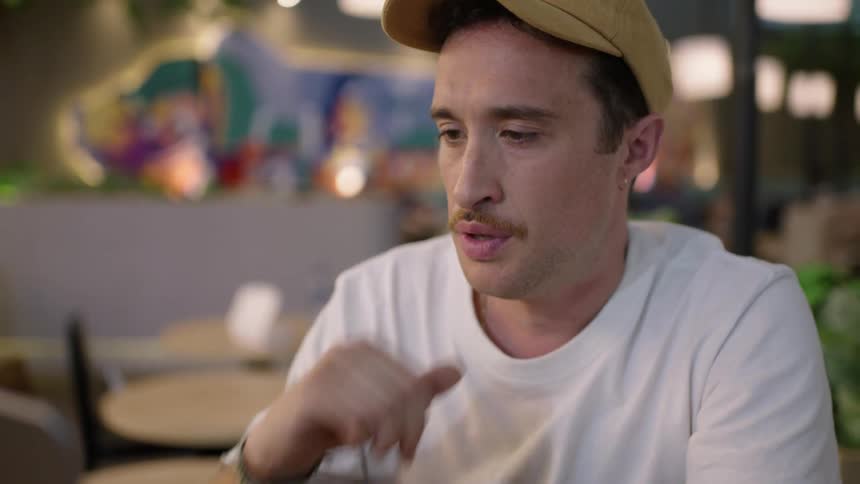 Man wearing yellow cap earrings mustache white shirt and black wristwatch uses laptop yawning at cafe