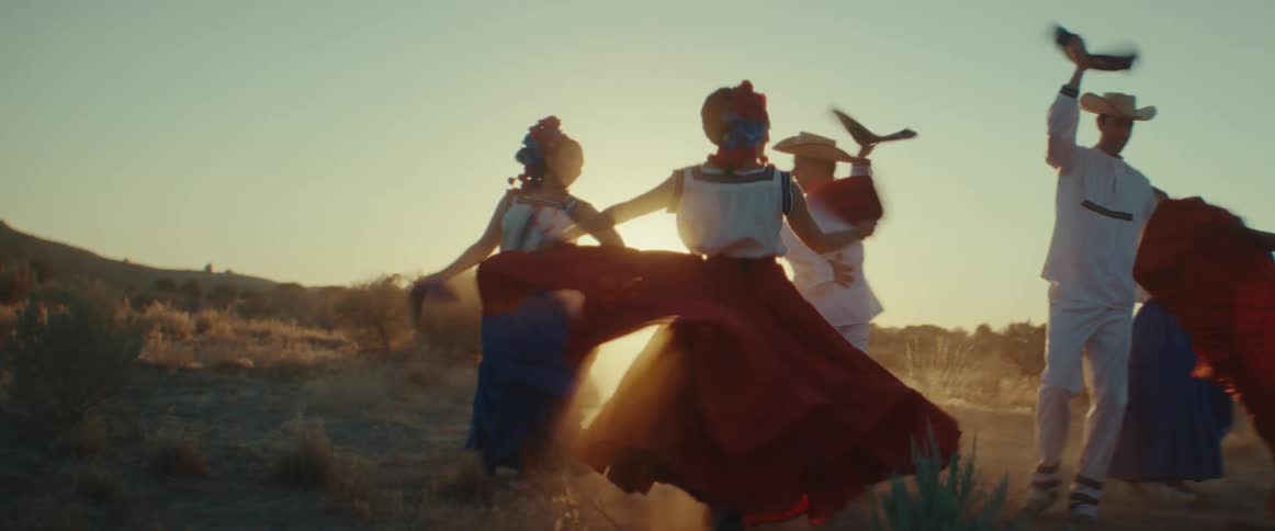 Male and female folk dancers wearing traditional costumes dance in field while backlit by sun