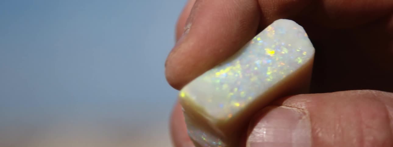 A person is holding a rectangular shaped opal that is white with rainbow speckles in their fingers