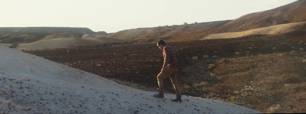 Man with plaid shirt and brown pants walks uphill in barren desert