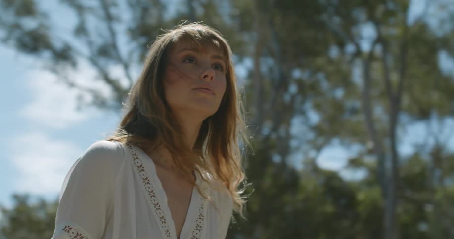A woman is walking outside looking around as her hair moves in the breeze and there is a forest of trees in the distance