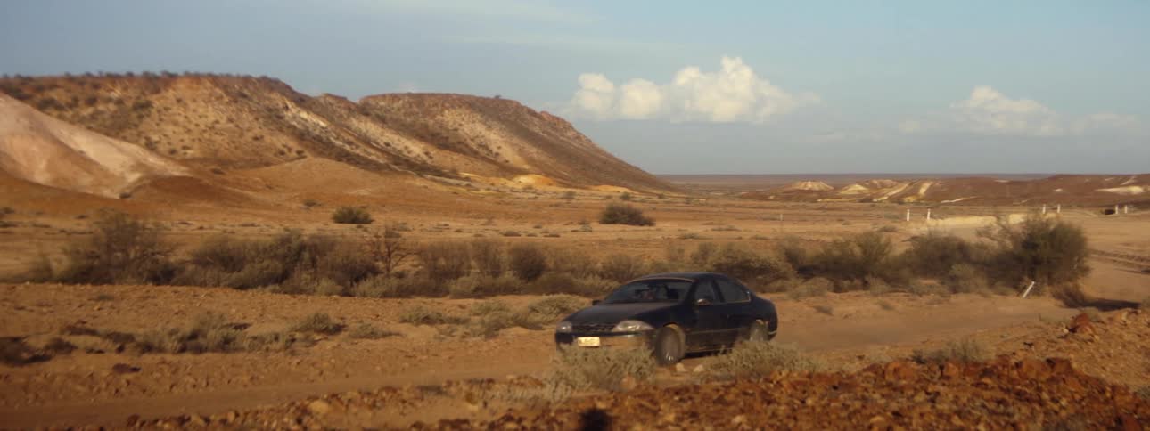 Car drives along dirt road through desert with scrub covered hills in the background