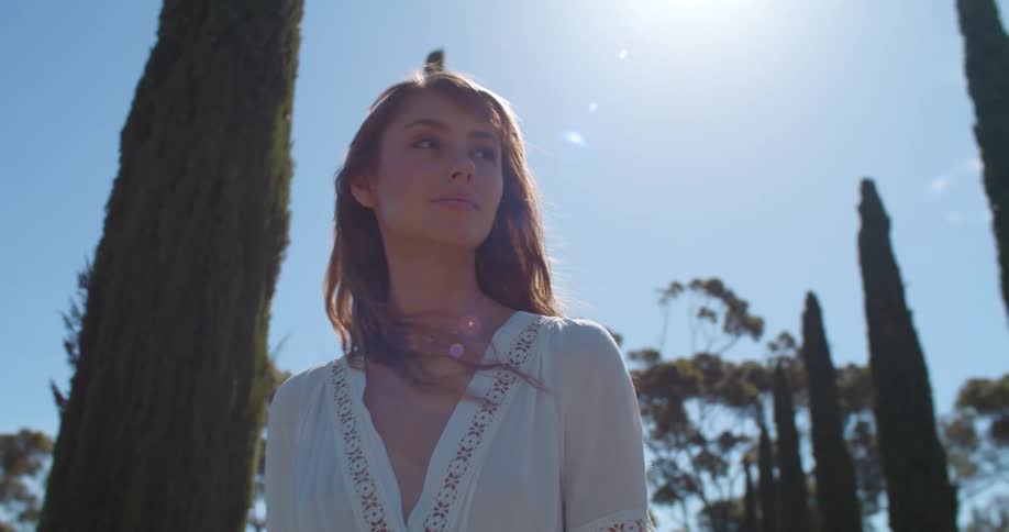 Woman with brown wavy hair and white dress walks in public park under shining sun