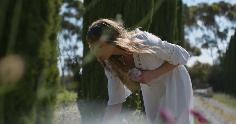 A young woman in a white dress leans over to pick flowers on a windy but sunny day
