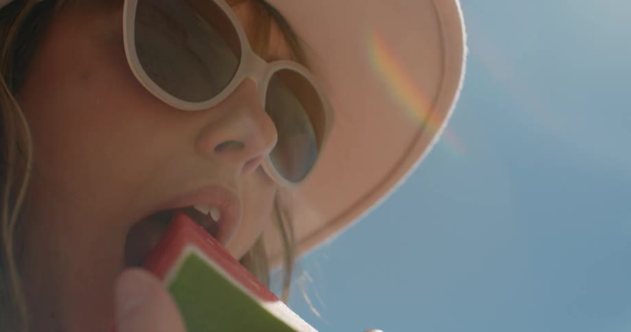 A woman wearing sunglasses takes a bite of a piece of watermelon on a sunny day