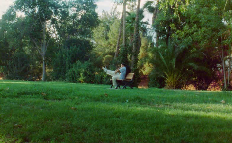 A woman is sitting on a park bench near trees doing a dance move and lying on the bench