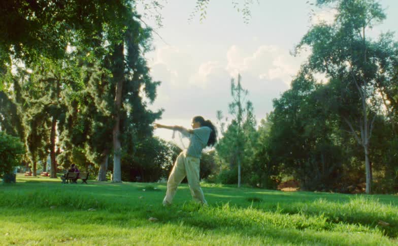 Woman dances in field in park with people sitting on bench in the background